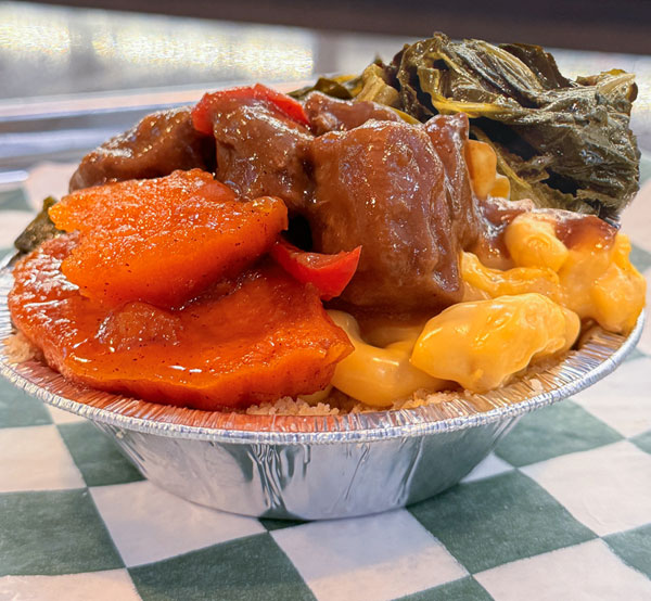 Hearty soul food bowl featuring collard greens, sweet yams, creamy mac & cheese, beef tips, and a cornbread crust.